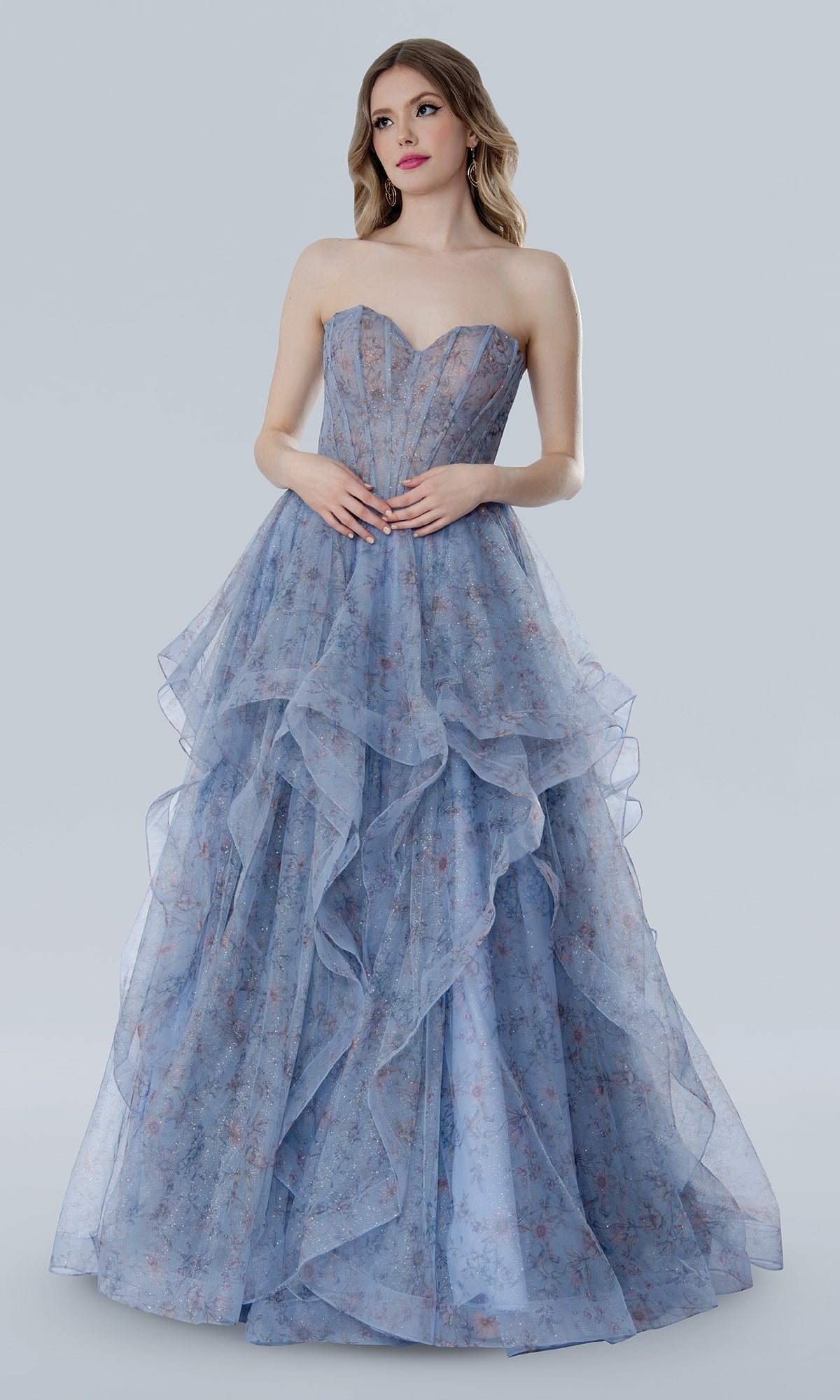 A woman models the Stella Couture 25119 Floral Prom Ball Gown, showcasing its strapless corset bodice and layered blue glitter tulle skirt with a subtle floral pattern, set against a plain light gray background.